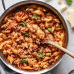 Close up shot of a pan filled with cooked pasta shells, ground turkey, and marinara on the gray background with a wooden spoon.