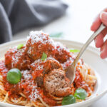 A plate of spaghetti topped with meatballs, tomato sauce, grated cheese, and fresh basil. A fork is cutting into a meatball.