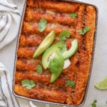 Baked enchiladas topped with melted cheese, sliced avocado, sour cream, and cilantro in a rectangular dish, with a halved avocado, lime wedges, and a striped towel nearby.