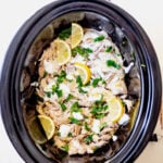 Overhead shot of shredded chicken breast garnished with lemon slices, parsley and yogurt sauce in a slow cooker.