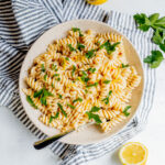 A bowl of rotini pasta garnished with parsley sits on a striped cloth, surrounded by lemon halves and parsley leaves, with a fork resting in the bowl.