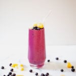 A straight on shot of a smoothie topped with blueberries and pineapple chunks in a clear glass. Blueberries and pineapple chunks are scattered on the marble surface around the glass.
