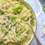 Overhead shot of cooked spaghetti with a green sauce topped with peas, basil leaves, and shredded parmesan cheese. A fork with spaghetti twisted around it lays on the edge of the white enamel pot.