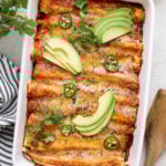 An overhead shot of a white rectangular baking pan with cooked enchiladas topped with enchilada sauce, melted cheese, avocado slices, jalapeño slices, and cilantro on a white background with a white and blue stripped napkin, wooden spoon, and a bunch of a cilantro.