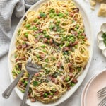 Overhead shot of a white oval dish with cooked spaghetti, cubed, ham, peas, and parmesan cheese. The dish is surrounded by a striped kitchen towel, a block of cheese, a plate with parsley, and a small wooden bowl with red pepper flakes.
