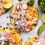 Overhead shot of salmon tacos with cabbage slaw and radishes. There are lime wedges, cilantro leaves, and a small wooden bowl with cilantro around it.