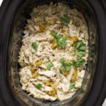 An overhead shot of shredded chicken topped with cilantro and green chilis in a black slow cooker.