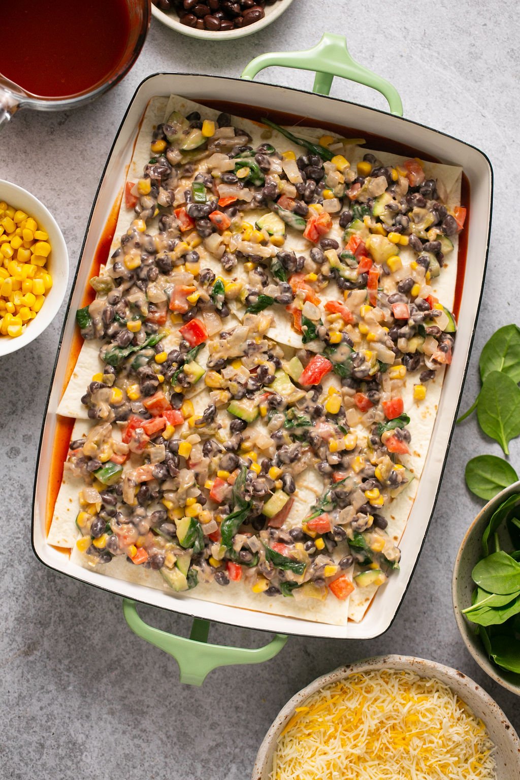 An overhead shot of a rectangular baking dish topped with tortilla halves and cooked veggies and surrounded by containers with spinach, shredded cheese, corn kernels, enchilada sauce, and black beans.