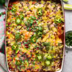 An overhead shot of a rectangular baking dish with a cooked vegetarian enchilada casserole. There are lime slices, sprigs of cilantro, and small bowls with jalapeños and cilantro around them.