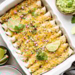 An overhead shot of a tablescape made up of a rectangular baking dish with enchiladas, an empty plate, a striped napkin, and small bowls with limes, cilantro, red onion, and guacamole.