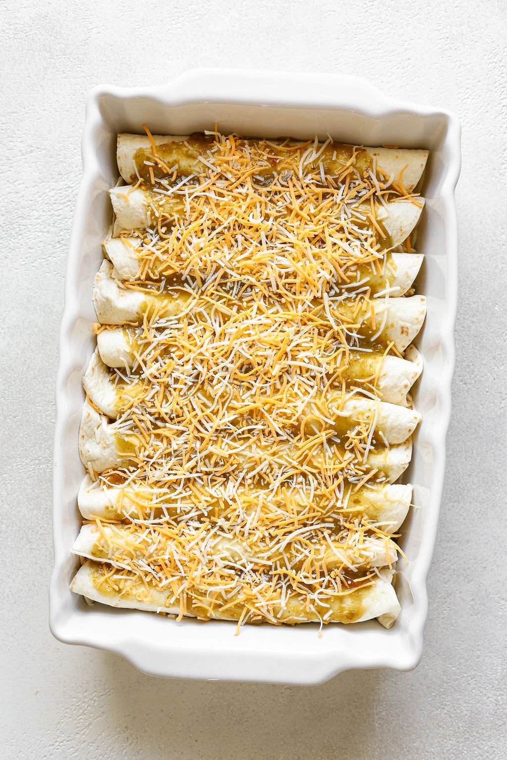 An overhead shot of a white rectangular baking dish with rolled enchiladas topped with salsa verde and shredded cheese.