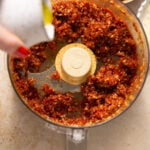An overhead shot of a food processor with a minced red paste. A hand is pouring olive oil into the paste.
