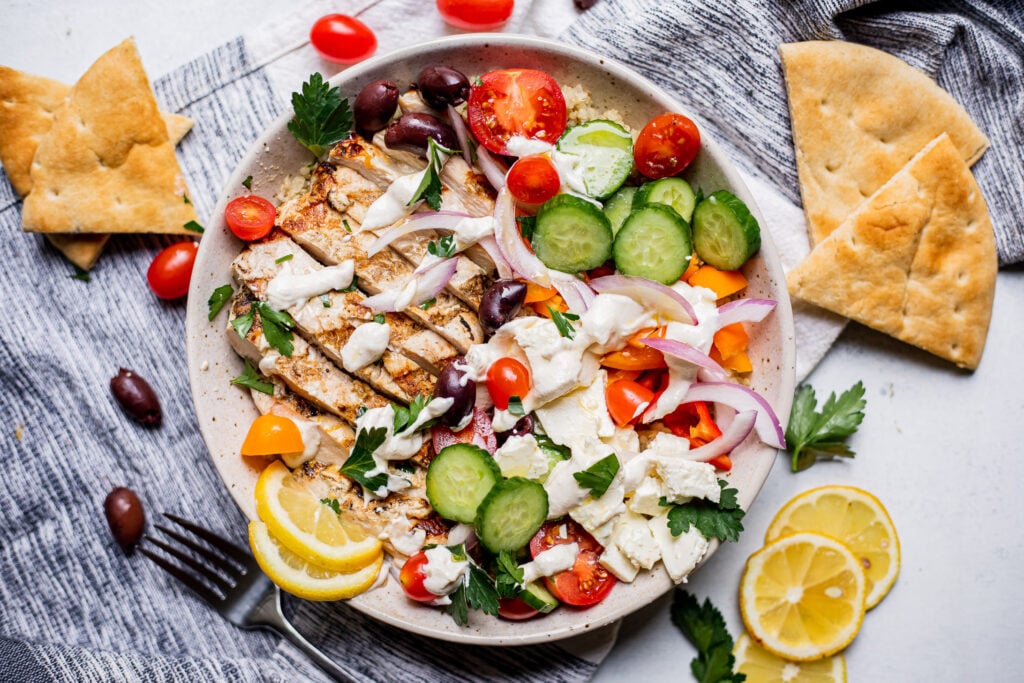 A bowl of Greek grilled chicken, tomatoes, cucumbers, red onion, feta cheese, olives, and pita bread, garnished with lemon slices and parsley.