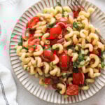 Overhead shot of a plate with pasta salad and a fork surrounded by glasses of water, tomatoes on the vine, a cloth napkin, glasses of water, and a small bowl with pepper.