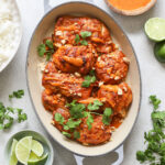 Overhead shot of an oval baking dish with cooked chicken thighs. The baking dish is surrounded by a lime cut in half, fresh herbs, and containers with sauce, lime wedges, and peanuts.