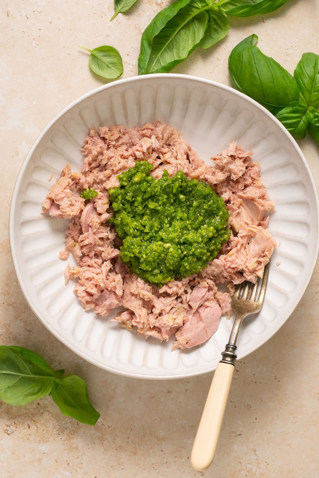 Overhead shot of a white plate with tuna topped with pesto and a fork. The plate is surrounded by basil leaves.
