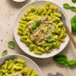Overhead shot of plates with pesto pasta topped with a tuna mixture. The plates are surrounded by basil leaves, a kitchen towel, and a small bowl with pesto.