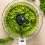Overhead shot of a food processor with pesto sauce surrounded by basil leaves.