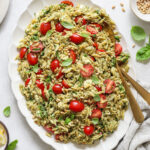 Overhead image of an oval platter with orzo pasta salad and two gold serving utensils sticking out of it.