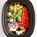 An overhead shot of a slow cooker with an assortment of raw vegetables and a sprig of basil.