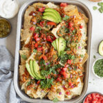 Overhead shot of a rectangular metal pan with nachos topped with tomatoes, jalapeños, avocado, and cilantro. The pan is surrounded by smaller bowls with sour cream jalapeños, avocado slices, chopped tomato, cilantro, and a dish towel.