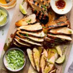 Overhead shot of a wooden cutting board with sliced grilled chicken, green onions, limes, soy marinade, and honey.