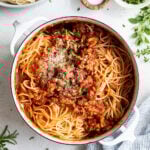 Overhead image of a white enameled dutch oven with spaghetti, with tomato sauce and ground turkey. The dutch oven is surrounded by a plate of cooked spaghetti, a bowl of parmesan cheese, a wooden bowl with salt, fresh herbs, and a white and blue striped tea towel.