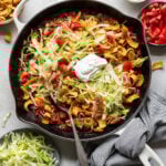 Overhead image of taco casserole topped with shredded lettuce, chopped tomatoes, and sour cream surrounded by containers with sour cream, tomatoes, Fritos, and shredded lettuce.