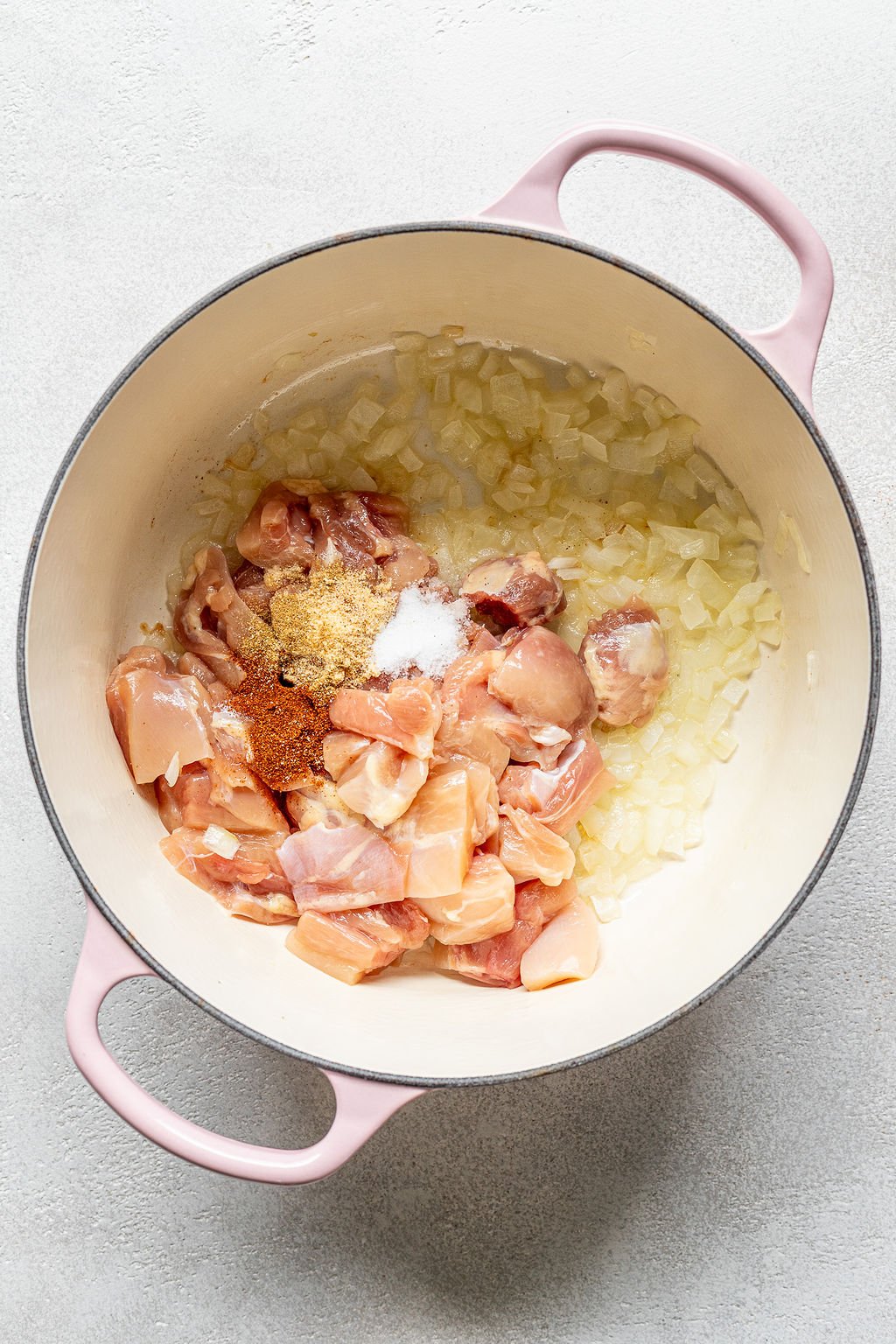 An overhead shot of an enameled cast iron pot with cooked onions and raw cubed chicken, and spices.