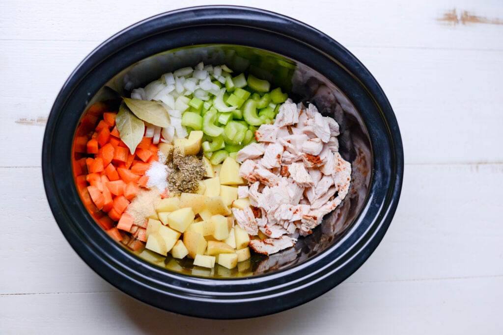 Overhead shot of cooked turkey, chopped potatoes, carrots, celery, onion and spices in a black slow cooker.