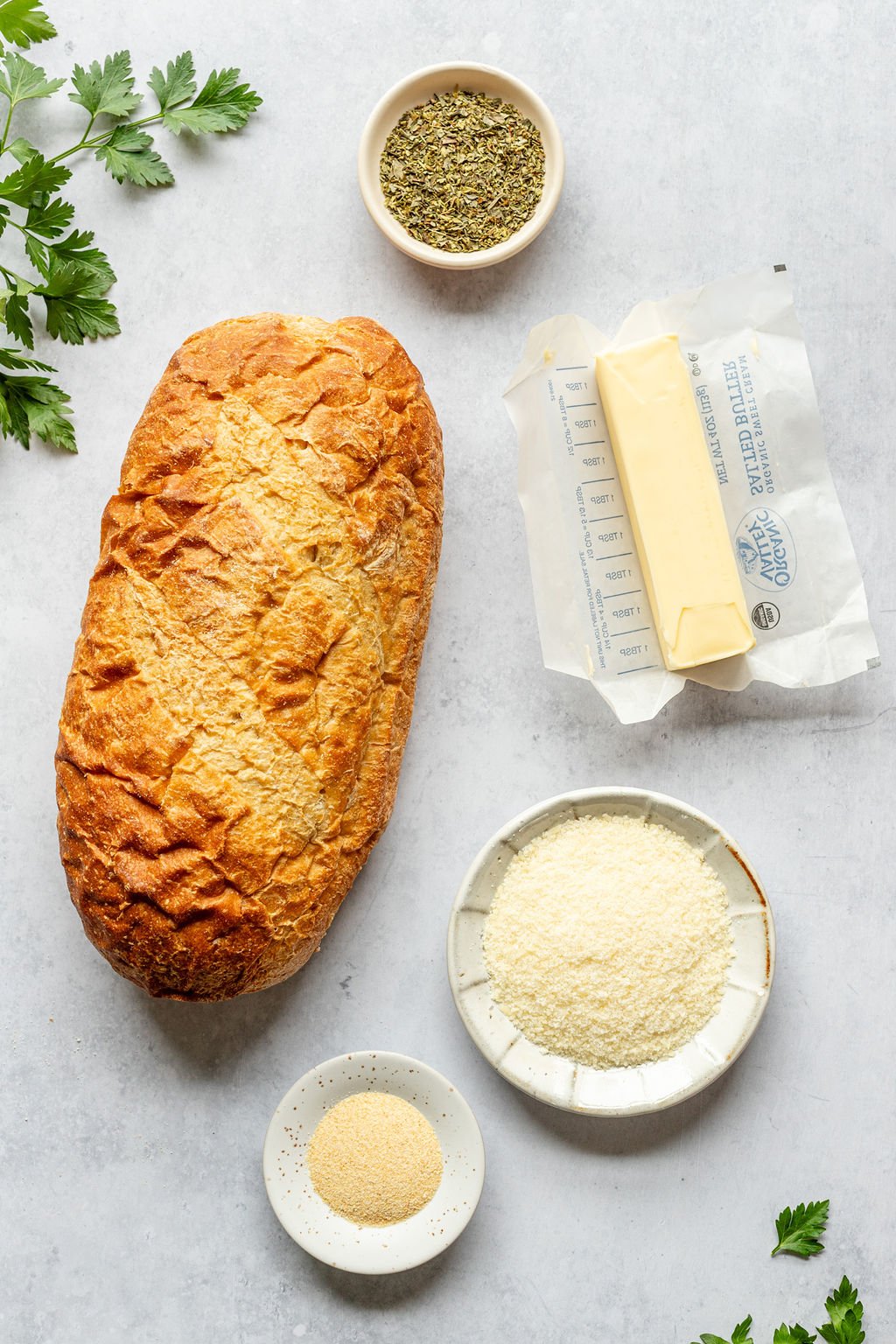 Overhead shot of the following ingredients: loaf of bread, stick of butter, spices, fresh parsley, parmesan cheese, and garlic powder.