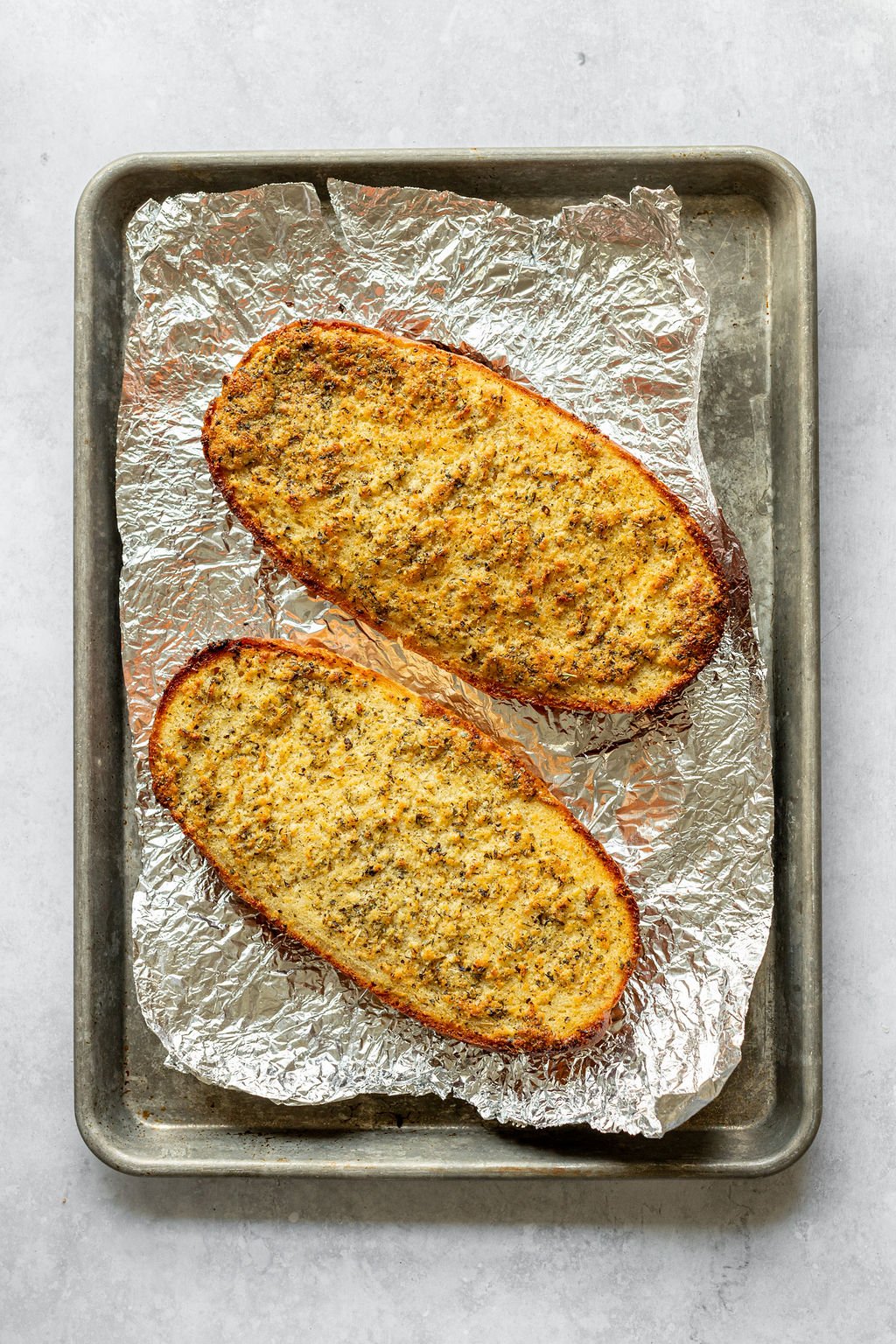 Overhead shot of two halves of a loaf of bread made into garlic bread on a baking sheet lined with foil.