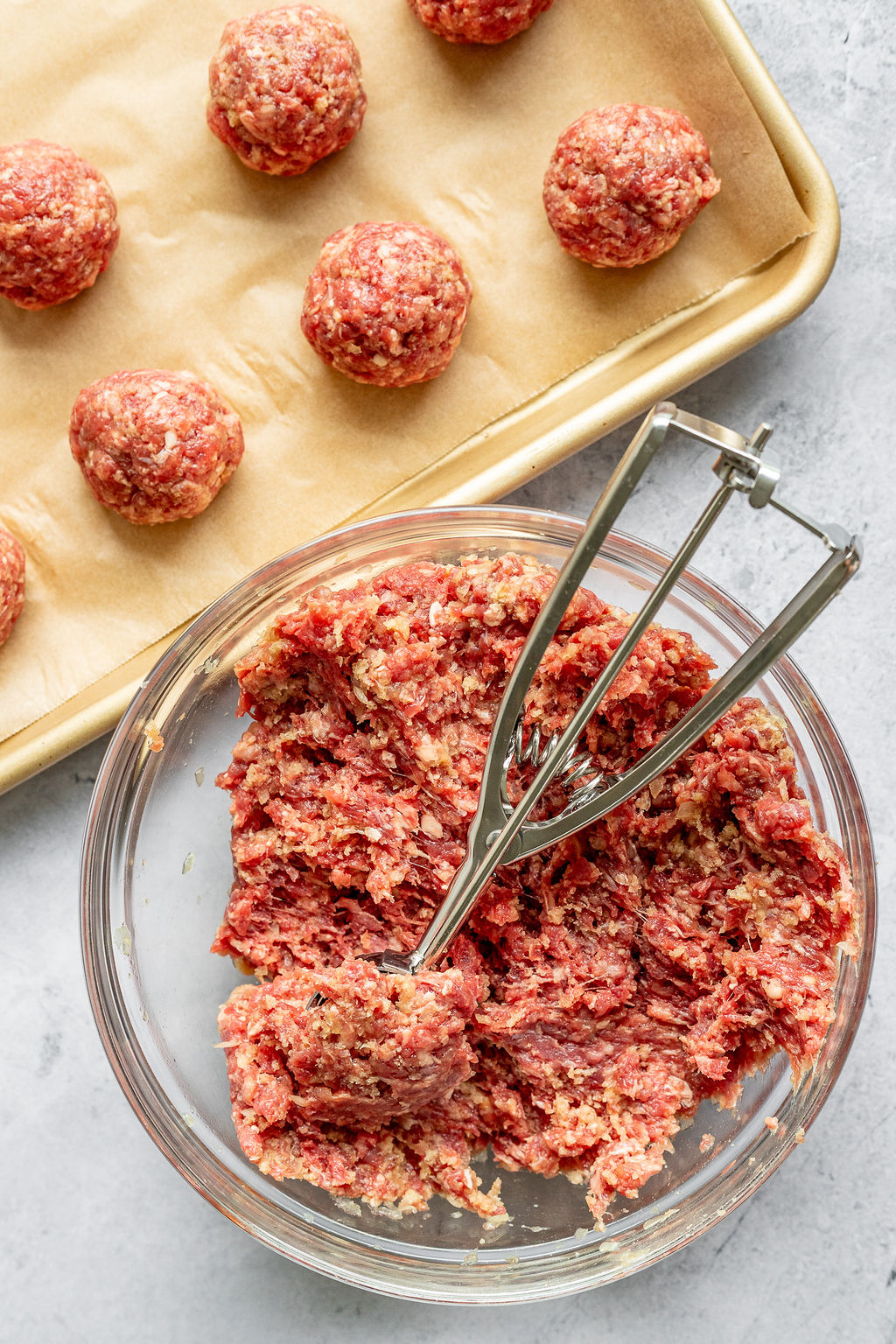 Overhead shot of meatballs being scooped by a cookie scoop and placed on a parchment lined baking sheet.