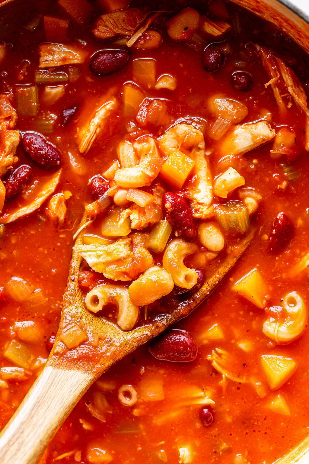 Closeup shot of a wooden spoon scooping beans, elbow pasta, shredded chicken, and vegetables out of a pot of soup.