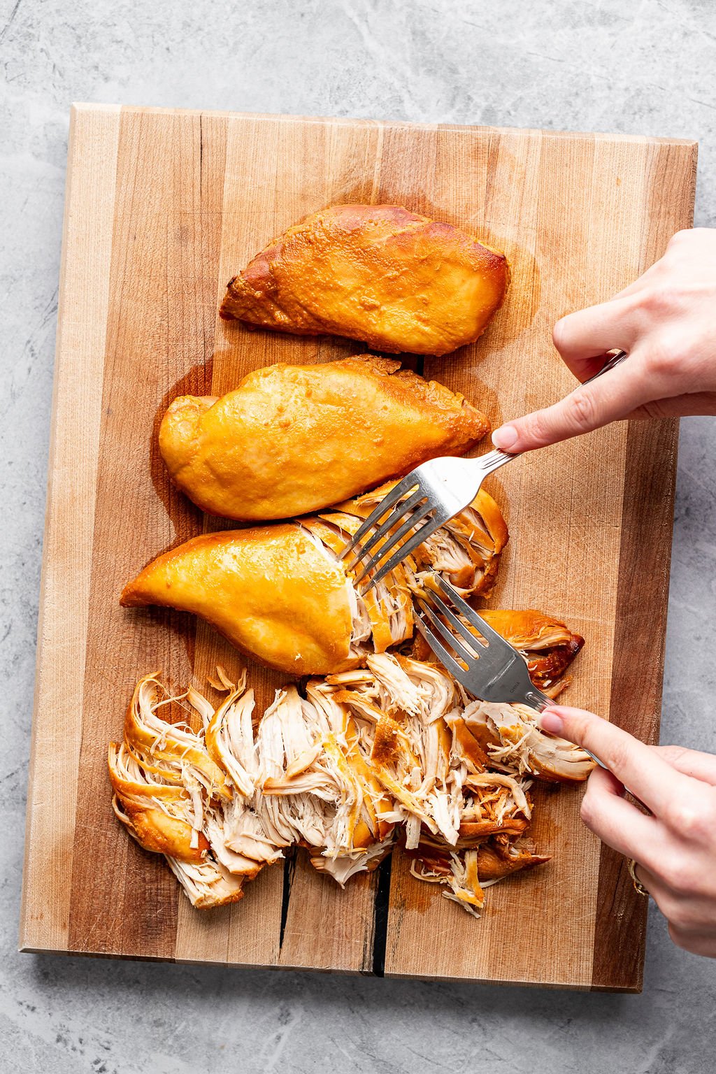 Overhead shot of chicken breasts being shredded with two forks on a wooden cutting board.