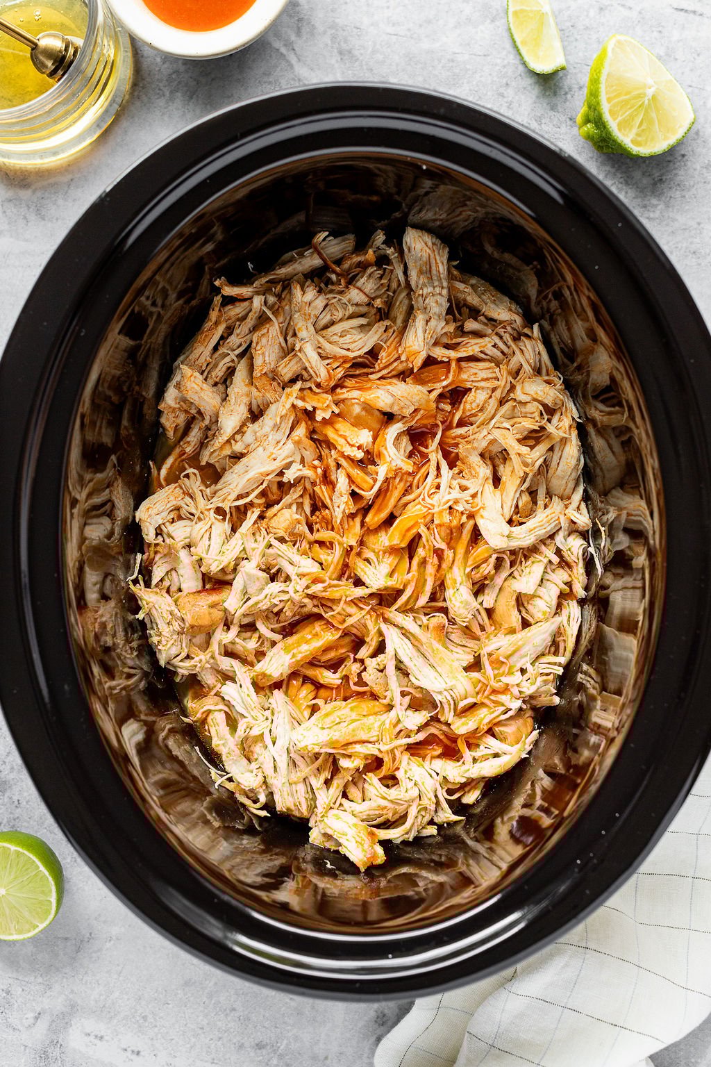 Overhead shot of shredded chicken in a slow cooker. The slow cooker is surrounded by honey, buffalo sauce, lime, and a tea towel.