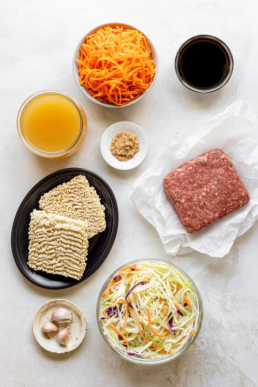Overhead shot of the following ingredients: ramen noodles, garlic cloves, cabbage slaw, raw ground beef, brown sugar, soy sauce, julienned carrots, and broth.