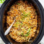 Overhead shot of ramen noodles in a slow cooker topped with sliced green onions and sesame seeds. A serving fork is sticking out of the noodles.