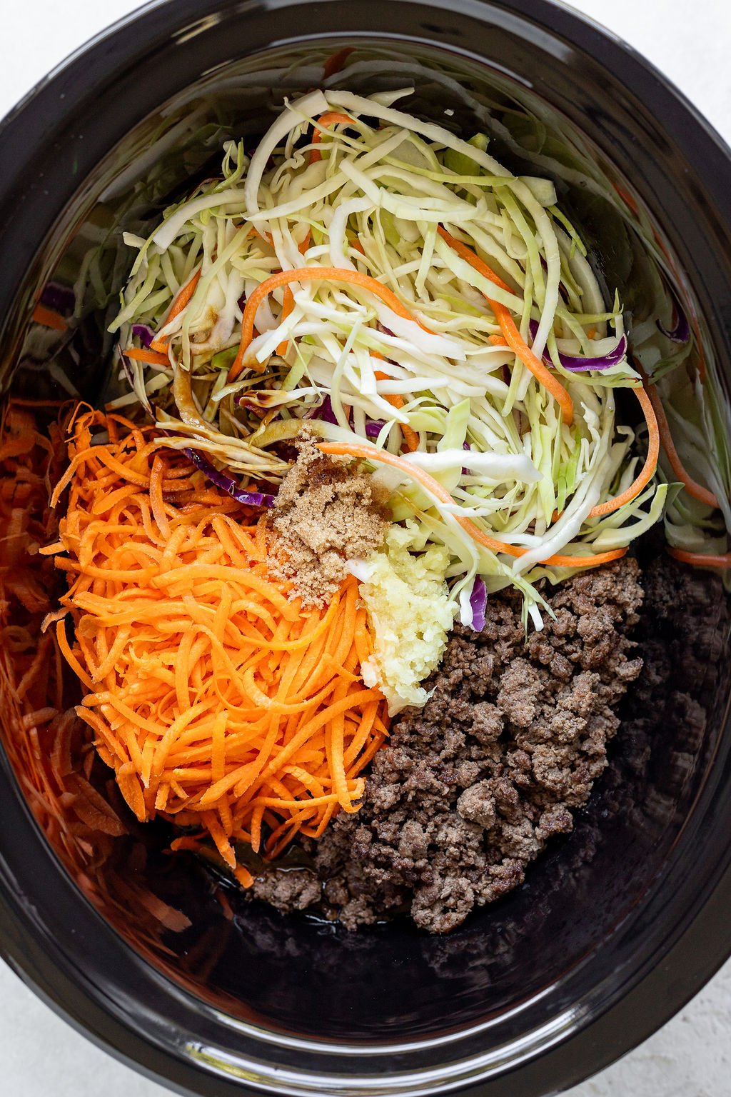Overhead shot of a slow cooker with julienned carrots, cabbage, brown sugar, garlic, and browned ground beef.