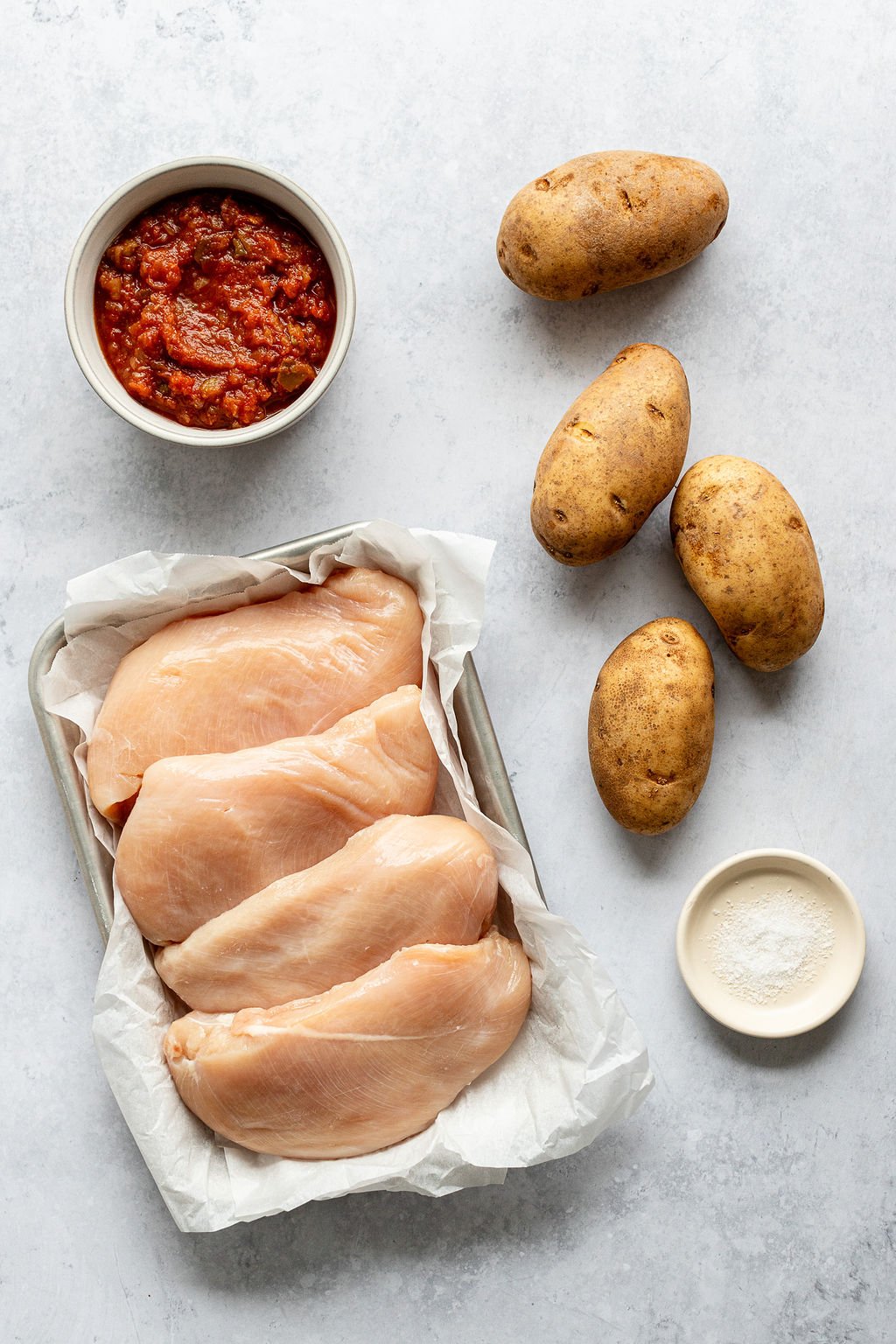 Overhead image of the following ingredients: potatoes, salsa, salt, and raw chicken breasts.