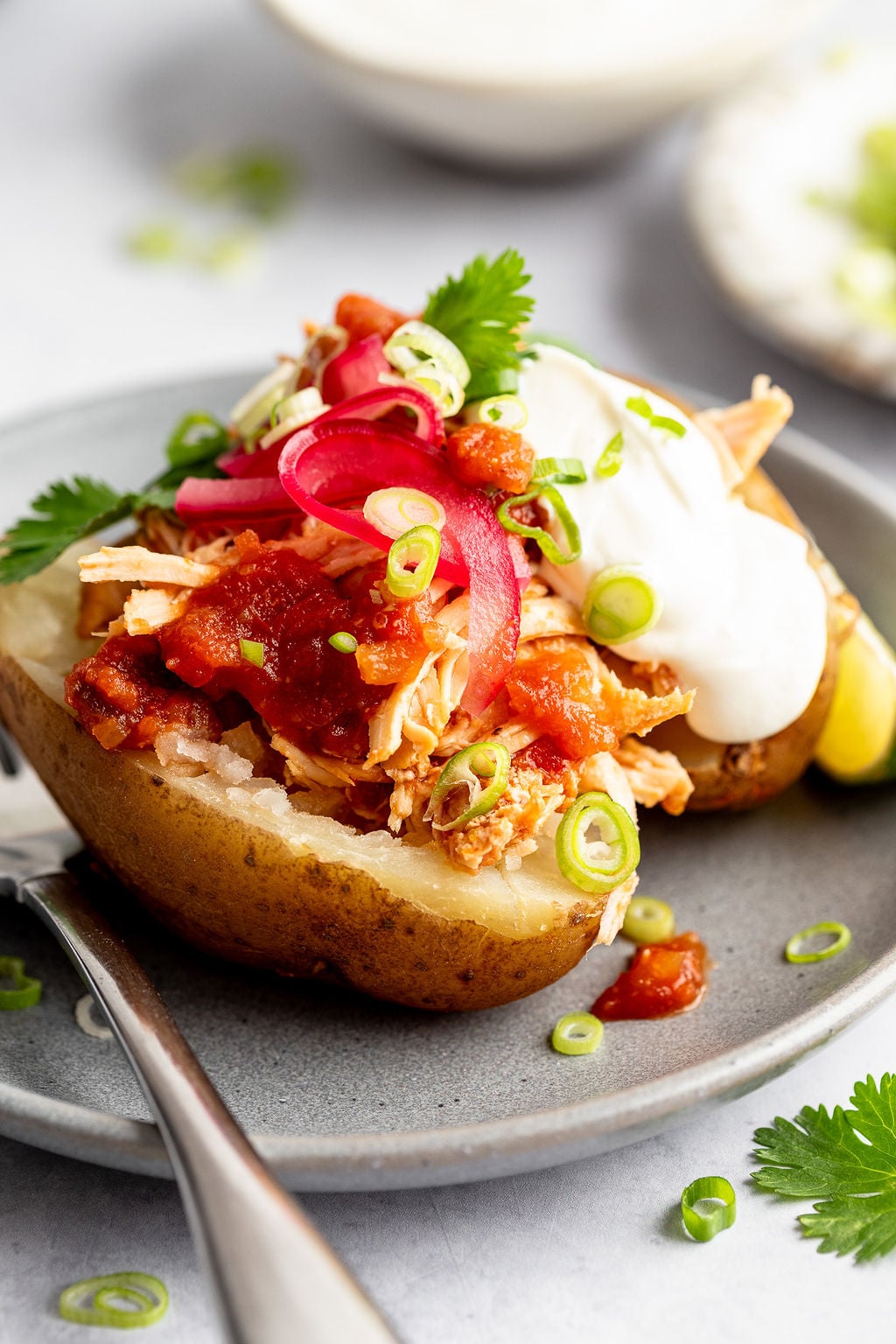 Close up image of grey plate with a fork and a baked potato stuffed with shredded chicken, salsa, scallions, sour cream, and cilantro.