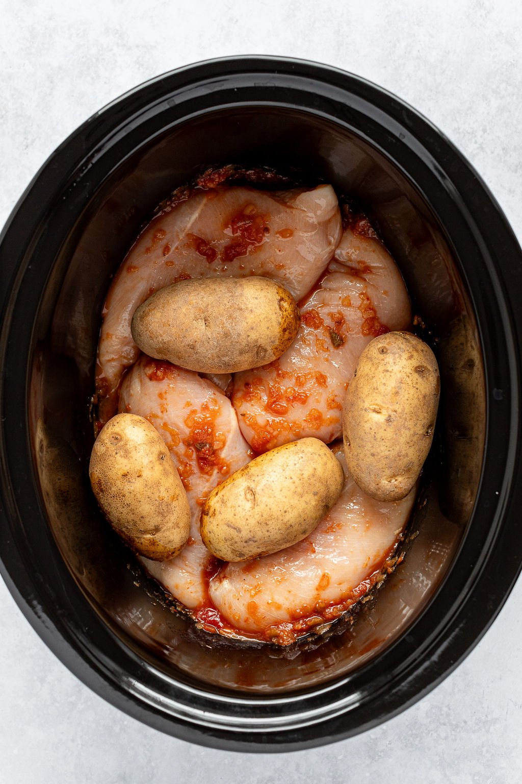Overhead shot of slow cooker with raw chicken breasts in red sauce and potatoes on top.