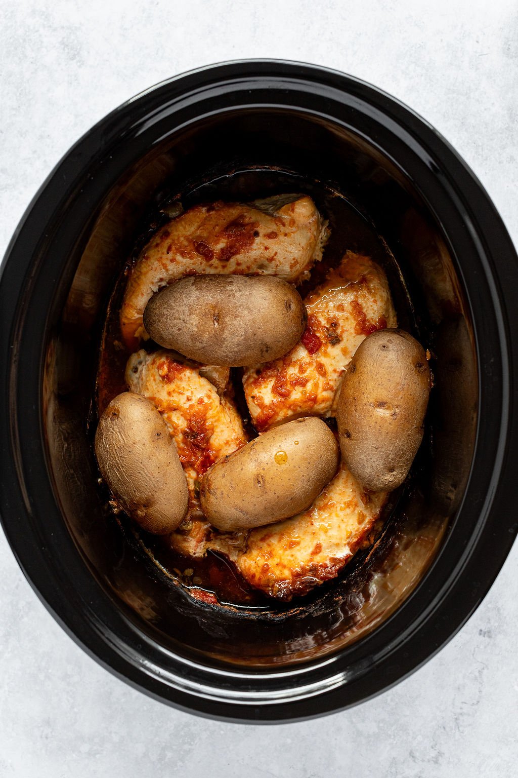 Overhead shot of slow cooker with cooked chicken in red sauce and potatoes on top.