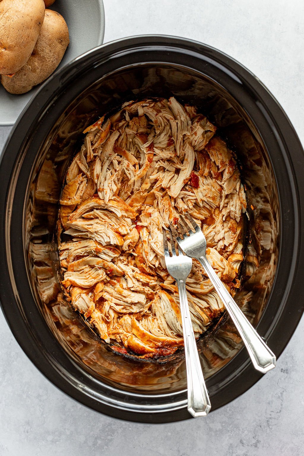 Overhead shot of slow cooker with two forks and shredded chicken in red sauce. Grey bowl with potatoes is off to the side.