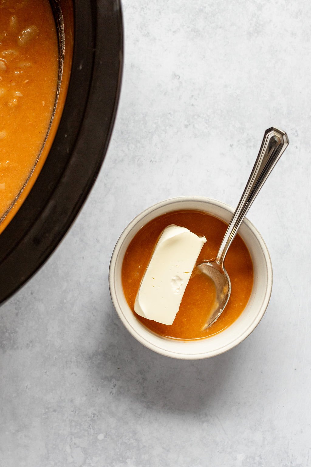 Overhead shot of a spoon in a small white bowl of buffalo chicken chili broth and a wedge of cream cheese. It is near a slow cooker with buffalo chicken chili.