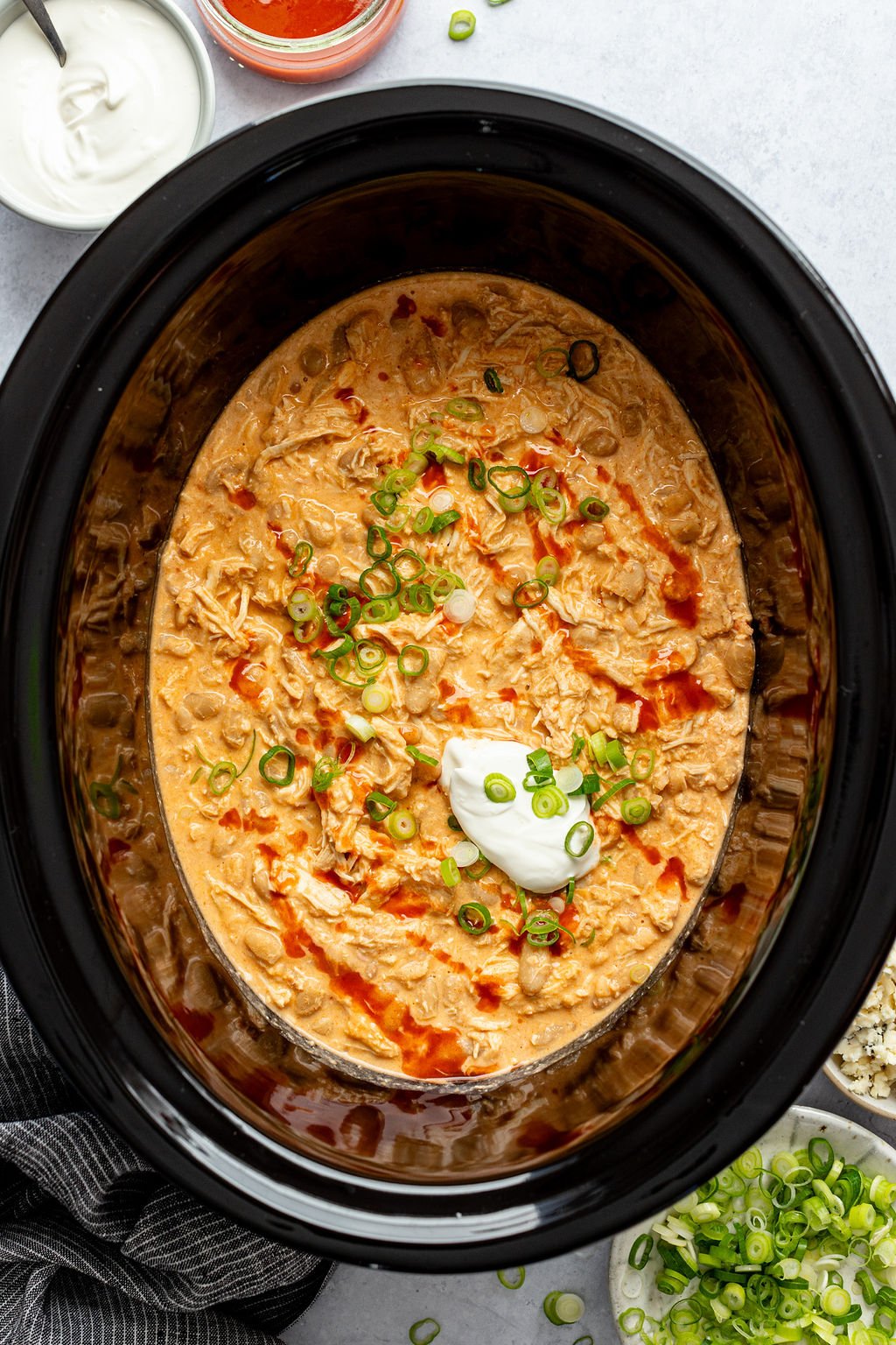 Close-up overhead shot of crock pot with cooked buffalo chicken chili topped with green onions, sour cream, and hot sauce. Crock pot is surrounded by a towel and small bowls of sour cream, hot sauce, green onions, and blue cheese.