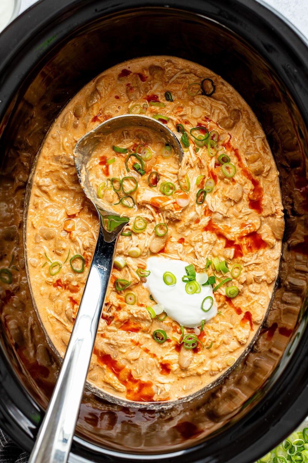 Close-up overhead shot of a ladle in a crock pot with cooked buffalo chicken chili topped with green onions, sour cream, and hot sauce.
