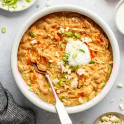 Close-up overhead shot of a white bowl with chicken chili topped with sour cream, hot sauce, and green onions. The bowl is surrounded by a towel and smaller dishes with blue cheese, sliced green onions, hot sauce, and sour cream.