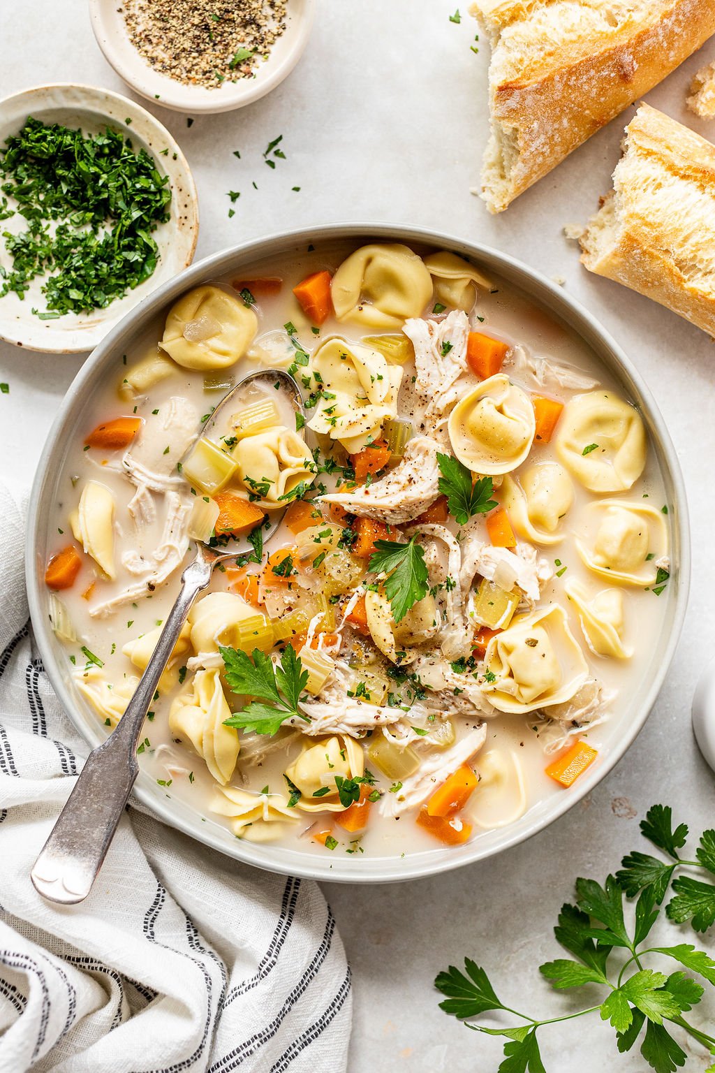 Overhead shot of a white bowl with creamy broth, diced carrots, shredded chicken, and tortellini, topped with fresh parsley and a hunk of baguette. Bowl is surrounded by a striped towel, sprigs of fresh parsley, baguettes, and small bowls of chopped parsley and pepper.