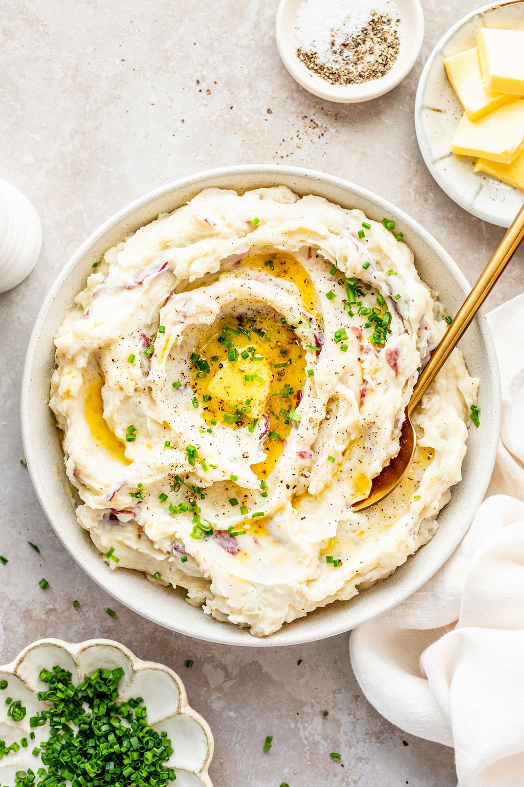 Overhead shot of serving mashed potatoes out of a white bowl. Bowl is surrounded by bowls of butter, salt and pepper, and chopped chives.